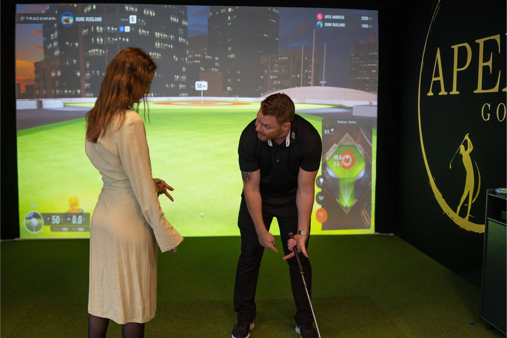 Apex Instruktør som viser en kvinne i kjole hvordan hun skal holde golfkøllen under en teambuilding. De står på en utslagsmatte i lokalene på Apex Golf Hinna Park.