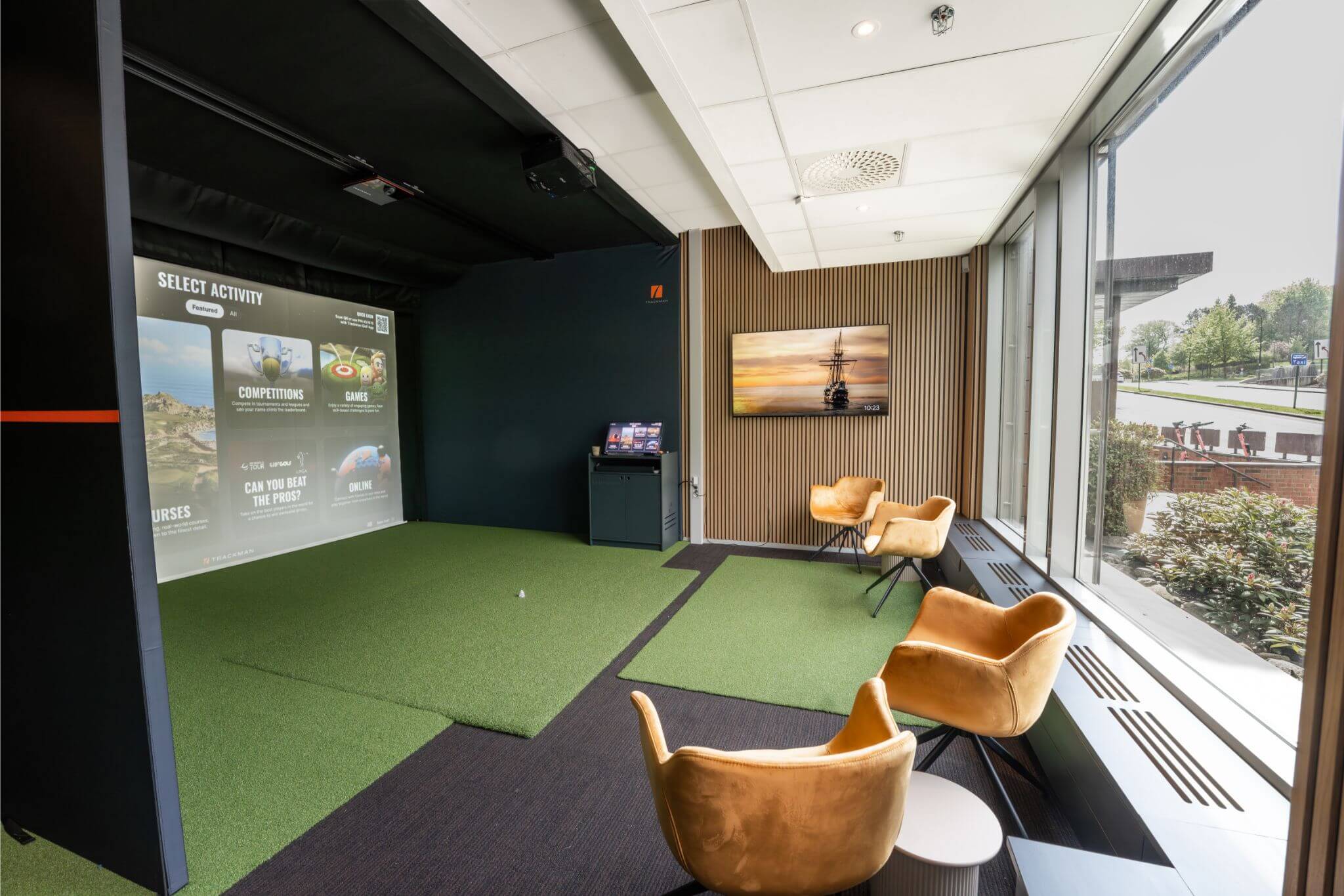 Den sørlige simulatorbayen hos Apex Golf Smedvigkvartalet, som viser utslagsmatten foran simulatorskjermen og et sitteområde med stoler på siden.